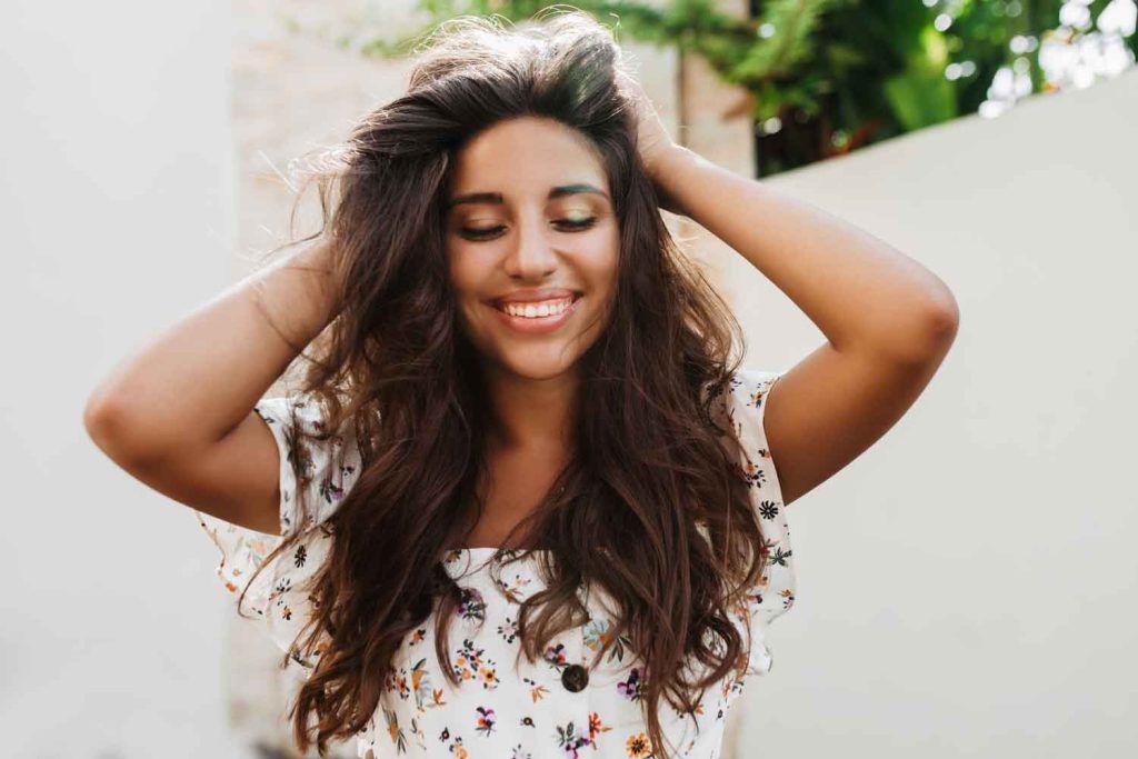 atractiva mujer bronceada blusa blanca con sonrisa encantadora posando pared blanca con plantas verdes