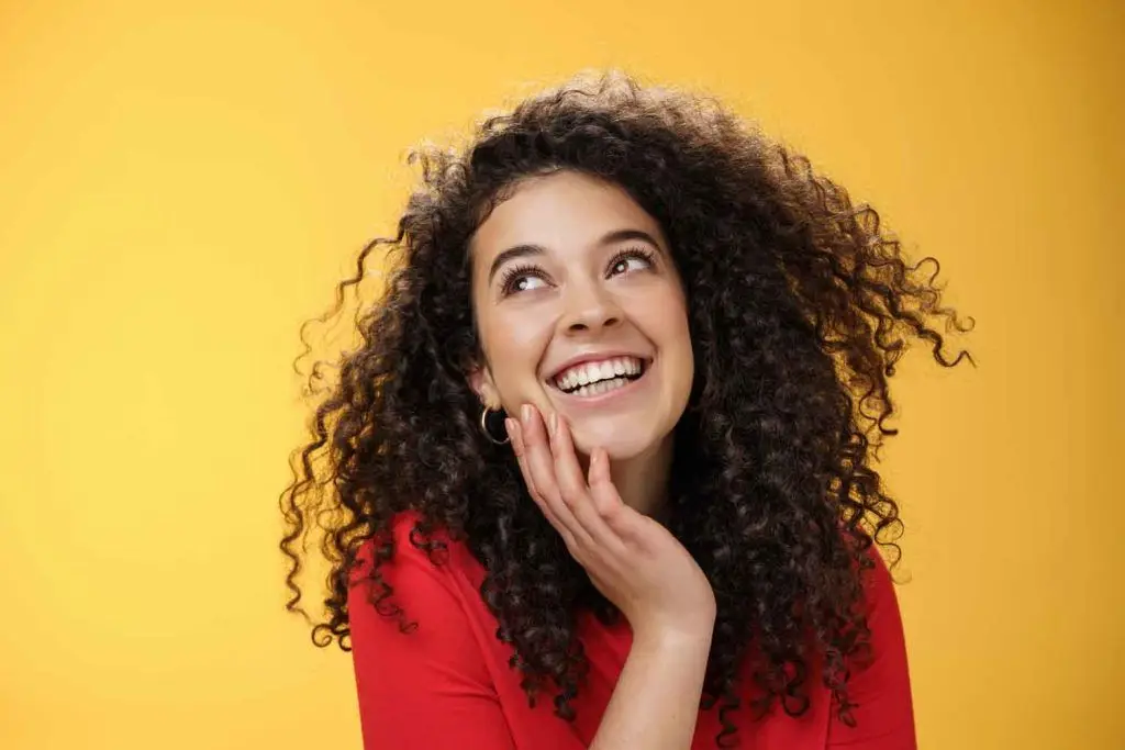 feliz positiva bonita joven con pelo rizado blusa roja riendo tonta despreocupada como mirando complacida esquina superior izquierda tocando la cara satisfecha encantada