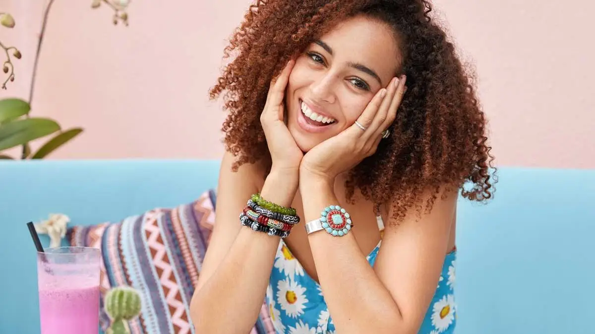 toma en interior mujer de raza mixta satisfecha con cabello rizado expresion alegre viste ropa de verano se sienta comoda en el sofa bebe un batido fresco