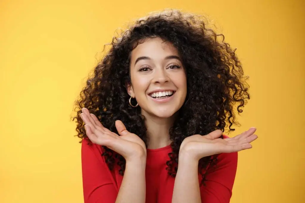 toma primer plano feliz amable tierna estudiante bastante caucasica pelo rizado piel perfecta sonriendo encantada sosteniendo palmas extendidas cerca cara divirtiendose sobre fondo amarillo